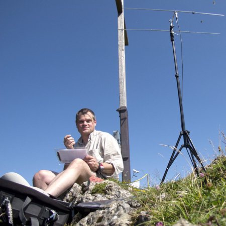 Olaf sitzt am Gipfelkreuz im Gras, schreibt ins Papierlog, daneben eine Vertikalantenne auf Stativ vor blauem Himmel